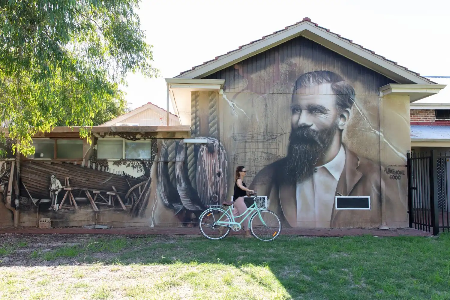 A person rides a bicycle past the Mandurah Museum, which features a large mural of historical scenes and a portrait on the building’s exterior.